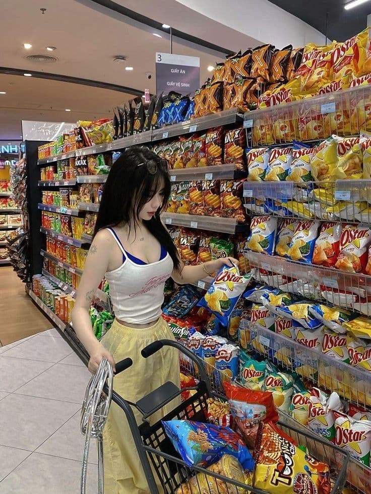 A woman in a white tank top and yellow pants picking out snacks from a colorful snack aisle in a grocery store