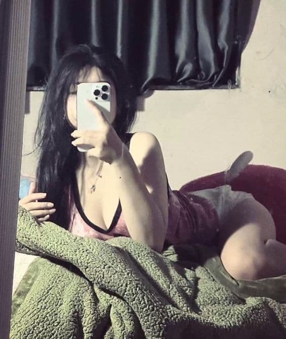 Mirror selfie of a woman with long dark hair holding an iPhone, wearing a pink top and light-colored shorts on a bed