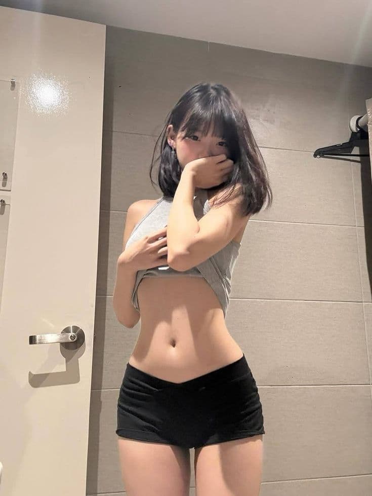 Young woman with short dark hair wearing a grey halter top and black shorts, posing in front of a white door