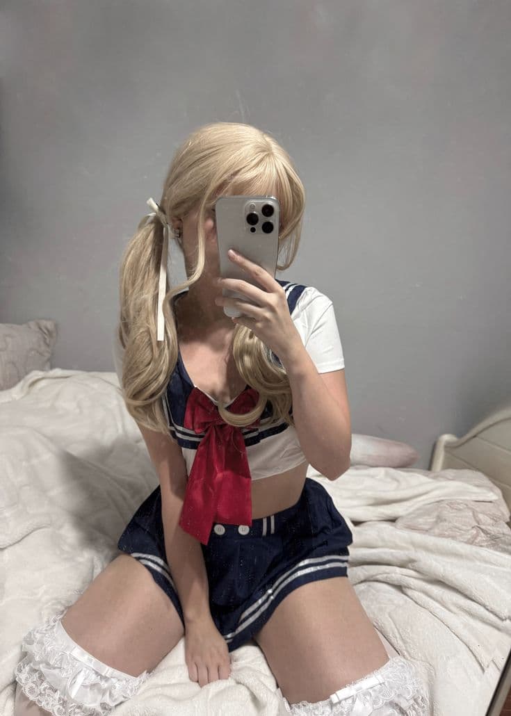Blonde girl in navy sailor outfit with red ribbon bow sitting on white bed