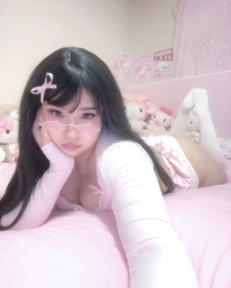 Asian girl in pink coquette outfit with heart glasses lying on pink bed surrounded by plushies
