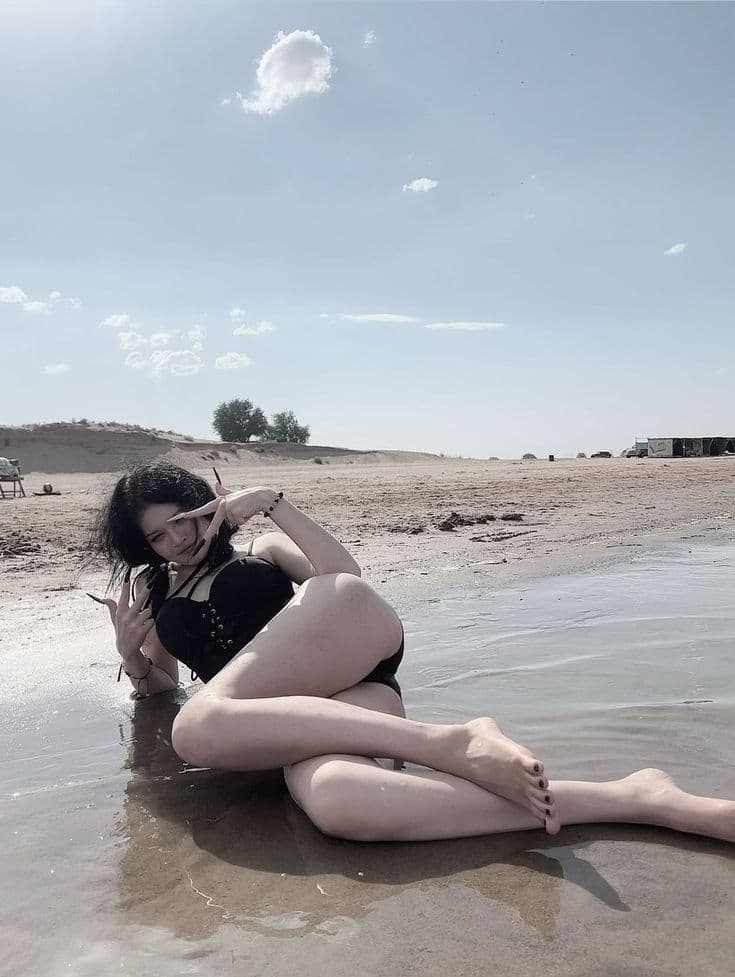 Young woman with dark hair reclining in shallow water on a sandy shore in a black lace-up bikini